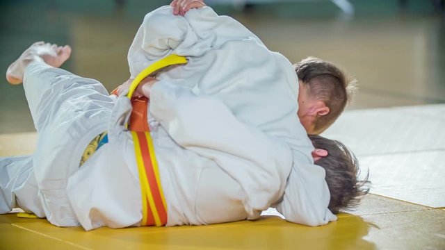 Kids are fighting on the floor during judo practise. They are wearing white judo uniforms and different colour of the belts.