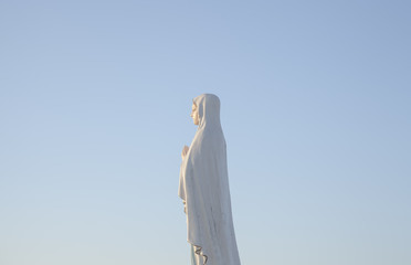 The statue of Our Lady Mother of Jesus Christ (Madonna, Mother Mary, Christian religion). Golden-hour clean sky in the background. Back view.
