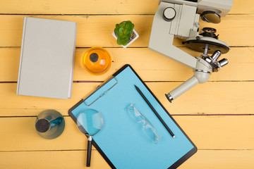 Education and science concept - microscope, book, magnifying glass, blank clipboard, eyeglasses and chemical liquids on the yellow desk