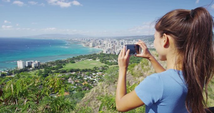 Young female hiker photographing Waikiki Beach and Honolulu through smartphone. Woman is looking at view from Diamond Head State Monument during summer vacation. She is wearing casual clothing.