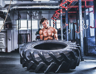 Muscular athletic bodybuilder flipping tire in cross gym