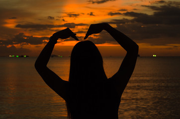 Silhouette of asian woman making sign of heart alone on beach with the sunlight when sunset.