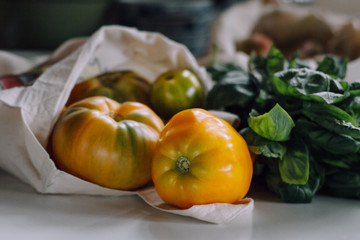 Eco tote bag with yellow tomatoes and bunch of basil on the kitchen table. Local food. Processed filtered image