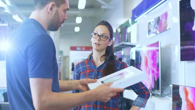 In the Electronics Store Professional Consultant Provides Expert Advice on Tablet Computer Specifications For Beautiful Young Woman. Store is Bright, Modern and Has all the Latest Devices. 4K UHD.