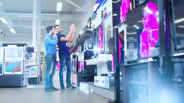 In the Electronics Store Professional Consultant Shows Latest 4K UHD TV's to a Young Man, They Talk about Specifications and What Model is Best for Young Man's Home. Store is Bright, Modern.