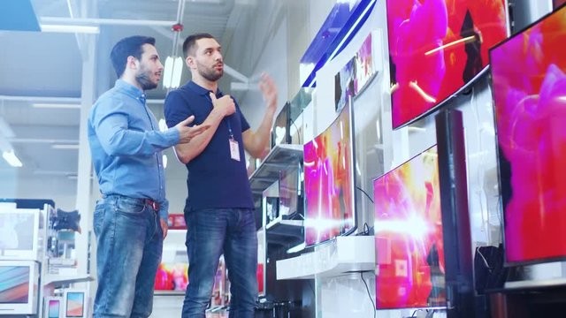 In the Electronics Store Professional Consultant Shows Latest 4K UHD TV's to a Young Man, They Talk about Specifications and What Model is Best for Young Man's Home. Store is Bright, Modern.