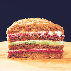 Close-up of sweet homemade layered honey cake on round wooden board on dark background, toned image. Cake made with different in color layer of sour cream