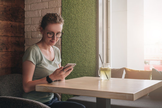 Hipster girl in glasses is sitting in cafe at table near window and looking on screen of smartphone. Businesswoman working online