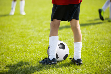 Close up legs and feet of football player in rwhite socks and black shoes playing with typical ball standing on green grass pitch outdoors. Child plays soccer training match