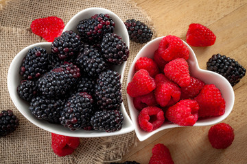 Heart healthy foods and good eating habits concept with two heart shaped white bowls of blueberries and raspberries with copy space. The raspberry bowl is smaller than the blackberry one