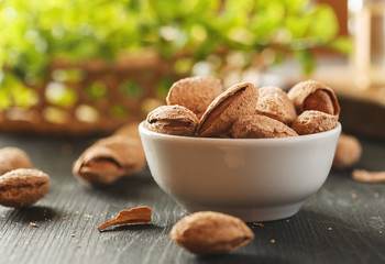 Almond in shell in a white ceramic bowl on a black wooden table with greenery behind it, closeup shot