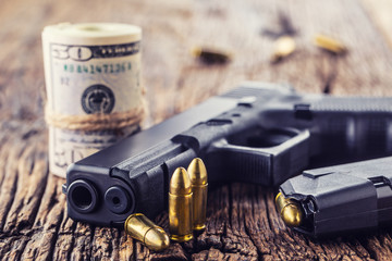 Obraz premium Gun and money. 9 mm pistol gun bullets strewn and roll dollar banknotes on rustic oak table
