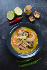 Plate of spicy green curry thai soup and some of its cooking ingredients on a dark metal background, above view