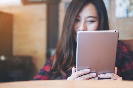 Closeup image of a smiley beautiful Asian woman holding ,using and looking at tablet pc sitting in modern cafe