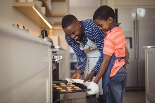 Father taking tray of fresh cookies out of oven with son in