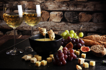 Gourmet Swiss fondue dinner on a winter evening with assorted cheeses on a board alongside a heated pot of cheese fondue with two forks dipping bread and white wine behind in a tavern or restaurant