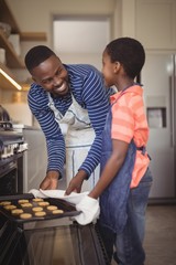 Father taking tray of fresh cookies out of oven with son in
