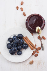 Homemade plum and chocolate jam served with fresh organic plums, cinnamon sticks and raw cocoa beans on white table