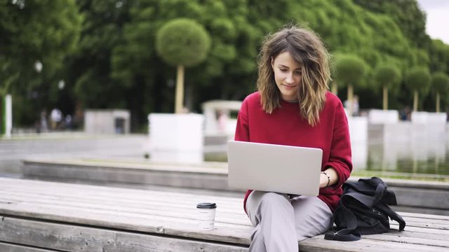 Young girl wearing a red sweatshirt and white pants is sitting near a fountain in a park and working at her laptop. Locked down real time medium shot
