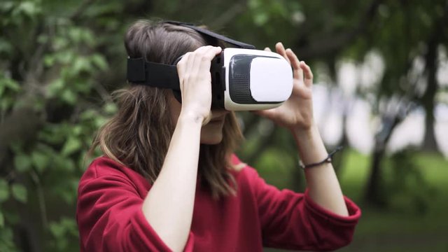 Side view of a young woman wearing a red sweater is using VR glasses while being at a park near large tree. Locked down real time close up shot