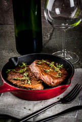 Lunch for one person. Homemade grilled beef steaks with thyme in a portioned frying pan, with a fork, knife and a glass of wine.  On black stone table, copy space