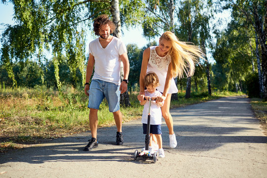 Happy Family In The Park. Parents With A Child On A Scooter Are Walking In Nature.