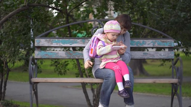 Mother with her little daughter dressed in a striped sweater together use the phone sitting on a bench in a public park on a cool evening. A young woman holds a baby girl on her lap.