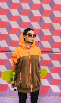 Portrait of trendy young female hip outfit with skate with colorful patterned wall