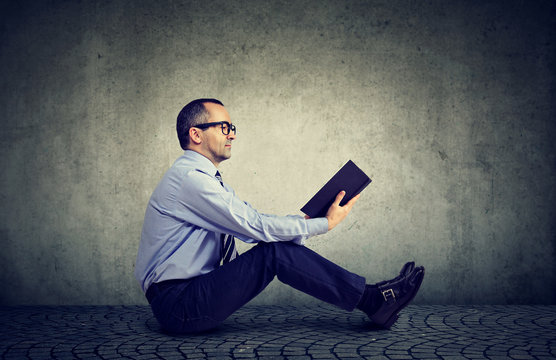 Man in glasses reading a book sitting on a floor
