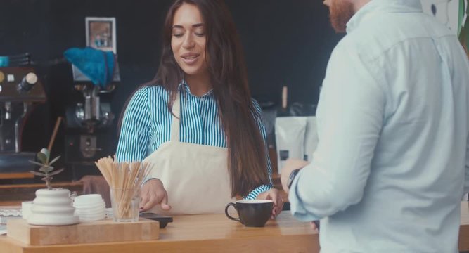 Attractive Caucasian female employee serving a coffee and taking mobile phone NFC payment from customer at modern coffee shop. 4K UHD 60 FPS

