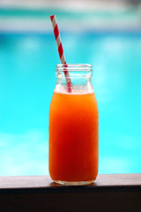 A carrot and orange juice in a glass bottle with a straw by a pool