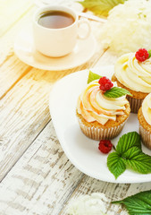 Delicate vanilla cupcakes with cream and raspberries on a white wooden background with a cup of coffee