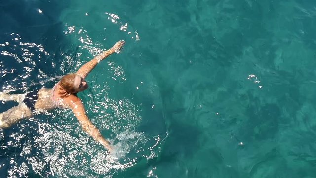 Young blonde woman dressed in black bikini suit swims breaststroke in the sparkling blue waves of the Aegean Sea on a sunny day in summer in slow motion