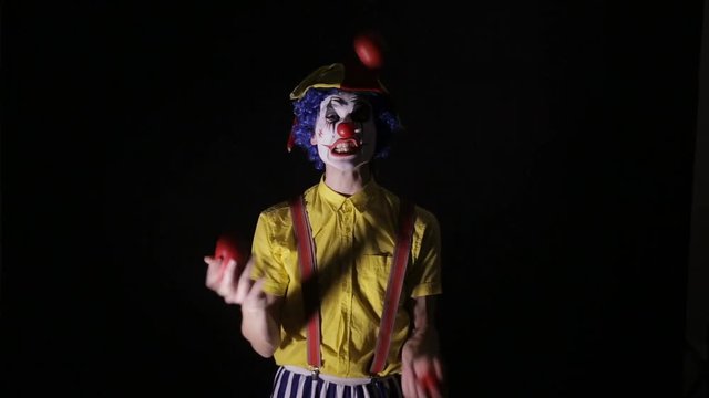 A clown juggles red apples and shows his teeth. 