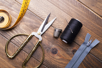 Scissors, tape measure, zipper, thread and needle next to thimble on wooden table.