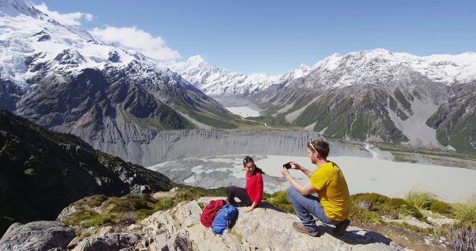 Travel vacation hiking couple taking photo in New Zealand by Mount Cook. People using smart phone taking picture on hike in Mount Cook national park landscape. Amazing nature.