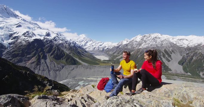 Hiking couple taking break eating food on alpine hike by Mount Cook New Zealand. Happy young people backpacking and tramping in Aoraki / Mt Cook National Park. Shot on RED EPIC.