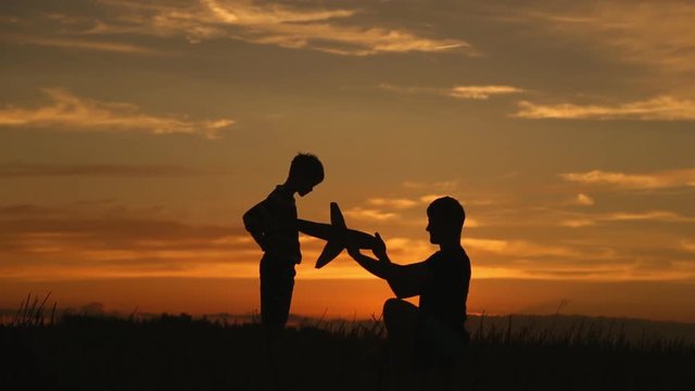A happy family. The boy on his father's shoulders is playing with an airplane toy. Silhouette at sunset by the lake.