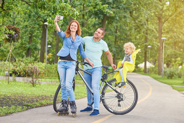 Happy active family taking a selfie with a smart phone while cycling and rollerblading together at the park copyspace.