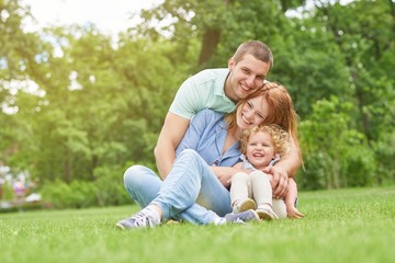 Naklejka na ściany i meble Happy young man smiling joyfully embracing his beautiful wife and daughter sitting on the grass together copyspace family love emotions weekend enjoyment affection parents marriage.