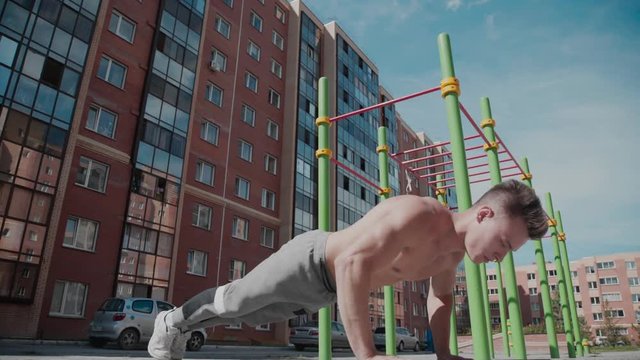 A man with a naked torso is engaged in a wright and presses. Young and athletic man doing sports outdoors and performs elements of workout. Training in courtyard. Street sports: exercises on bar.