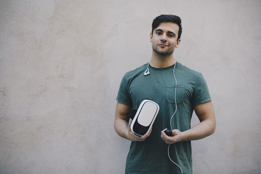 Portrait of computer programmer holding VR glasses and remote control against beige wall in office
