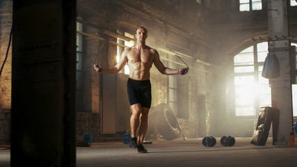 Obraz na płótnie Canvas Muscular Fit Man Exercises with Jump / Skipping Rope in a Deserted Factory Hardcore Gym. He's Sweaty from His Cross Fitness Exhausting Training.
