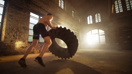 Naklejka na ściany i meble Strong Muscular Man Lifts Tire as Part of His Cross Fitness Program. He's Covered in Sweat and Works out in a Abandoned Factory Remodeled into Gym.