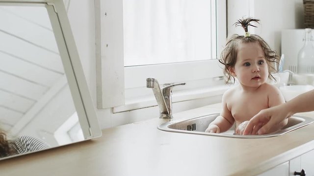 Baby taking bath in kitchen sink. Child playing in sunny bathroom with window. Little boy bathing. Water fun for kids. Hygiene and skin care for children. Bath room interior