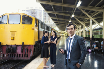 Business man hold mobile stand at railway with couple women and yellow train in background.