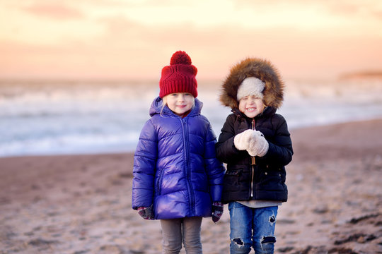Two cute little sisters having fun together at winter beach on cold winter day. Kids playing by the ocean.