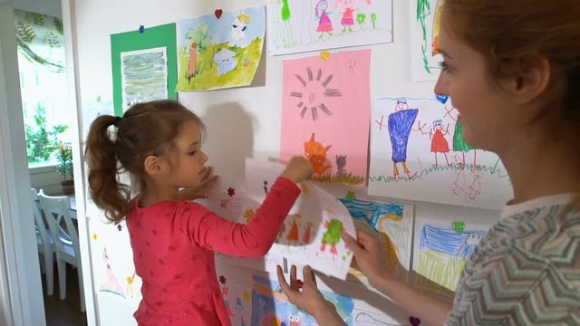 Little cute girl and her mother hang children's drawings on the wall in the nursery. Slow motion.