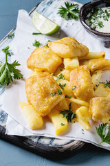 Traditional british fast food fish and chips. Served with white cheese sauce, lime, parsley, on white paper and black serving board over blue concrete background. Close up