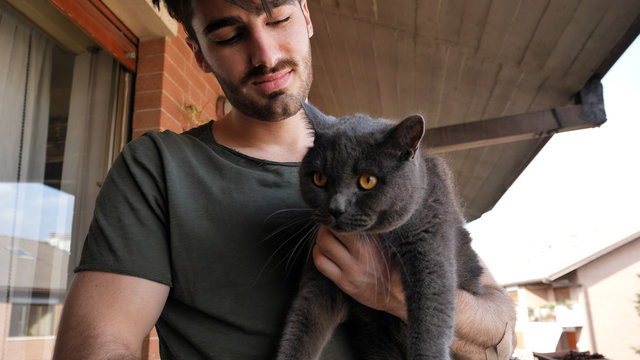 Handsome Young Animal-Lover Man Outside The House, Holding, Hugging And Petting His Gray Domestic Cat Pet.